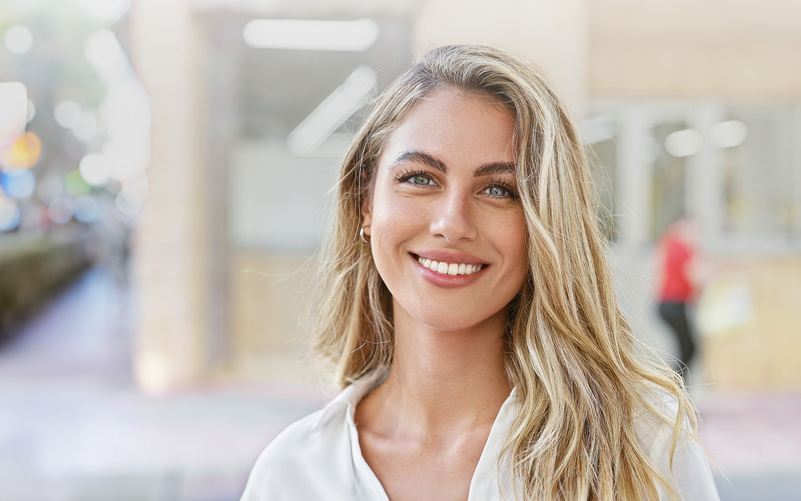 A woman with long blonde hair stands outdoors, smiling at the camera, wearing a white top and earrings, against a blurred background of a city street.