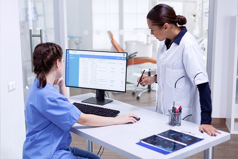 A professional healthcare setting with two individuals a woman in scrubs working at a desk with a computer monitor displaying a webpage, and another woman standing behind her, both engaged in their tasks within a modern office environment.