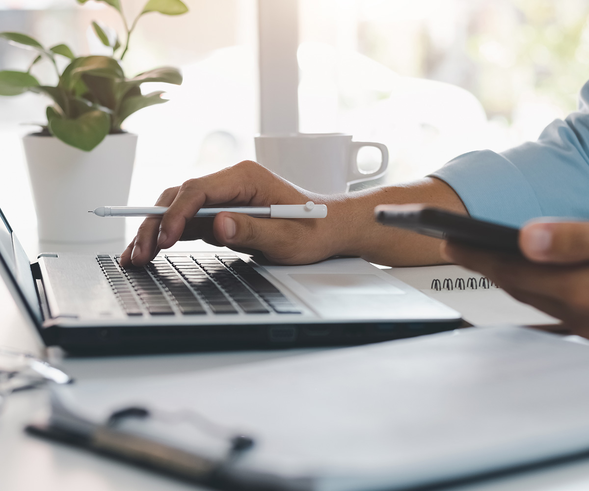 A person using a laptop at a desk with a pen and phone nearby, set against an indoor office background.